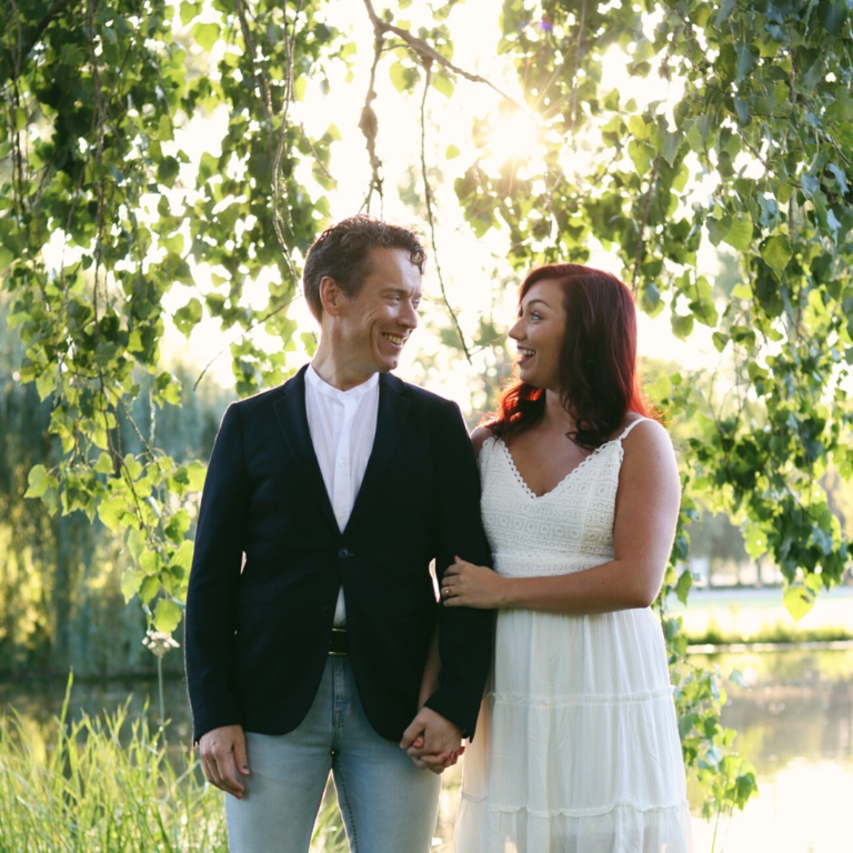 Greg and Brianna arm-in-arm outside with water and a willow tree behind them